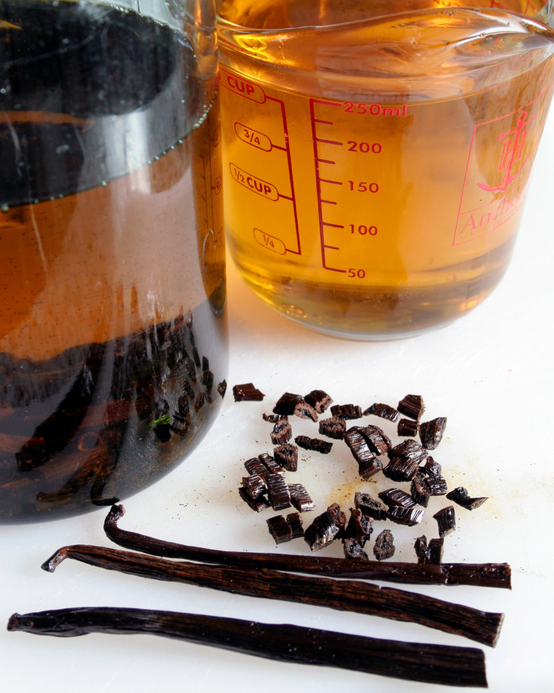 A close-up of Berry Beautiful’s vanilla infusion process. A Pyrex measuring cup holds filtered Red Raspberry Seed Oil and Cranberry Seed Oil infused with Tahitian and Madagascar vanilla beans, showing its deep golden color. To the left, a dark glass bottle contains the unfiltered infusion. In the foreground, whole and chopped vanilla beans rest on a white surface, highlighting the rich, aromatic ingredients used in the maceration process.