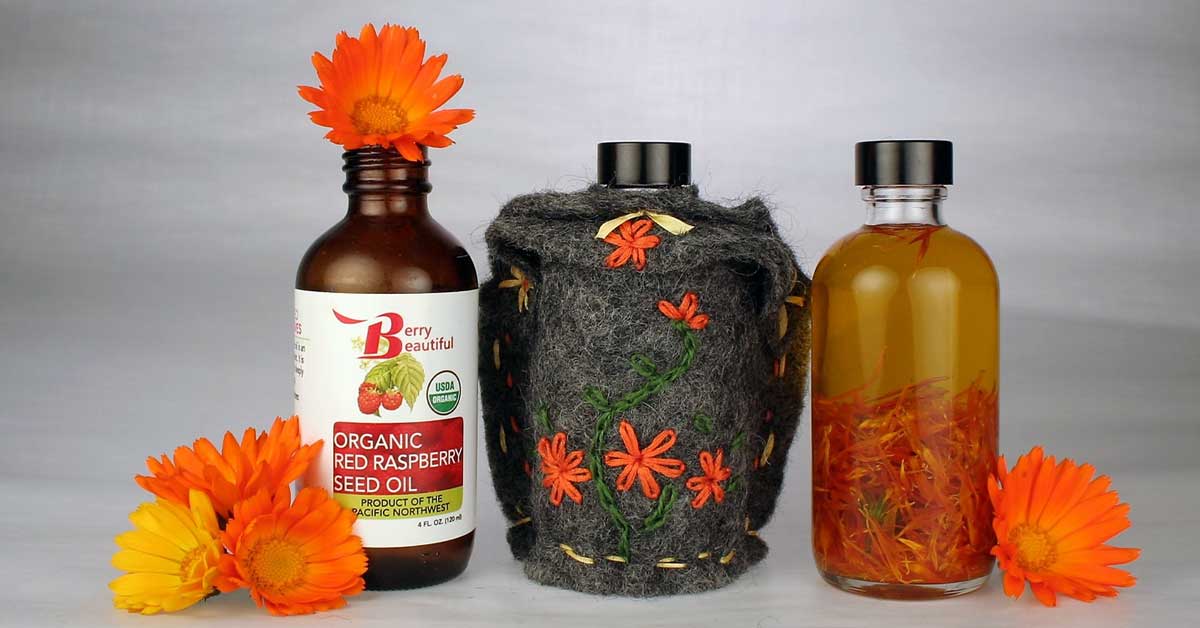 Three bottles of organic raspberry seed oil. The bottle on the left has a label that says "Beautiful Organic Red Raspberry Seed Oil". The bottle in the middle has a felt cover with embroidered designs. The bottle on the right is clear and shows raspberry seed oil with calendula petals infusing in the bottle.  This image shows that the oil can be used for infusions.